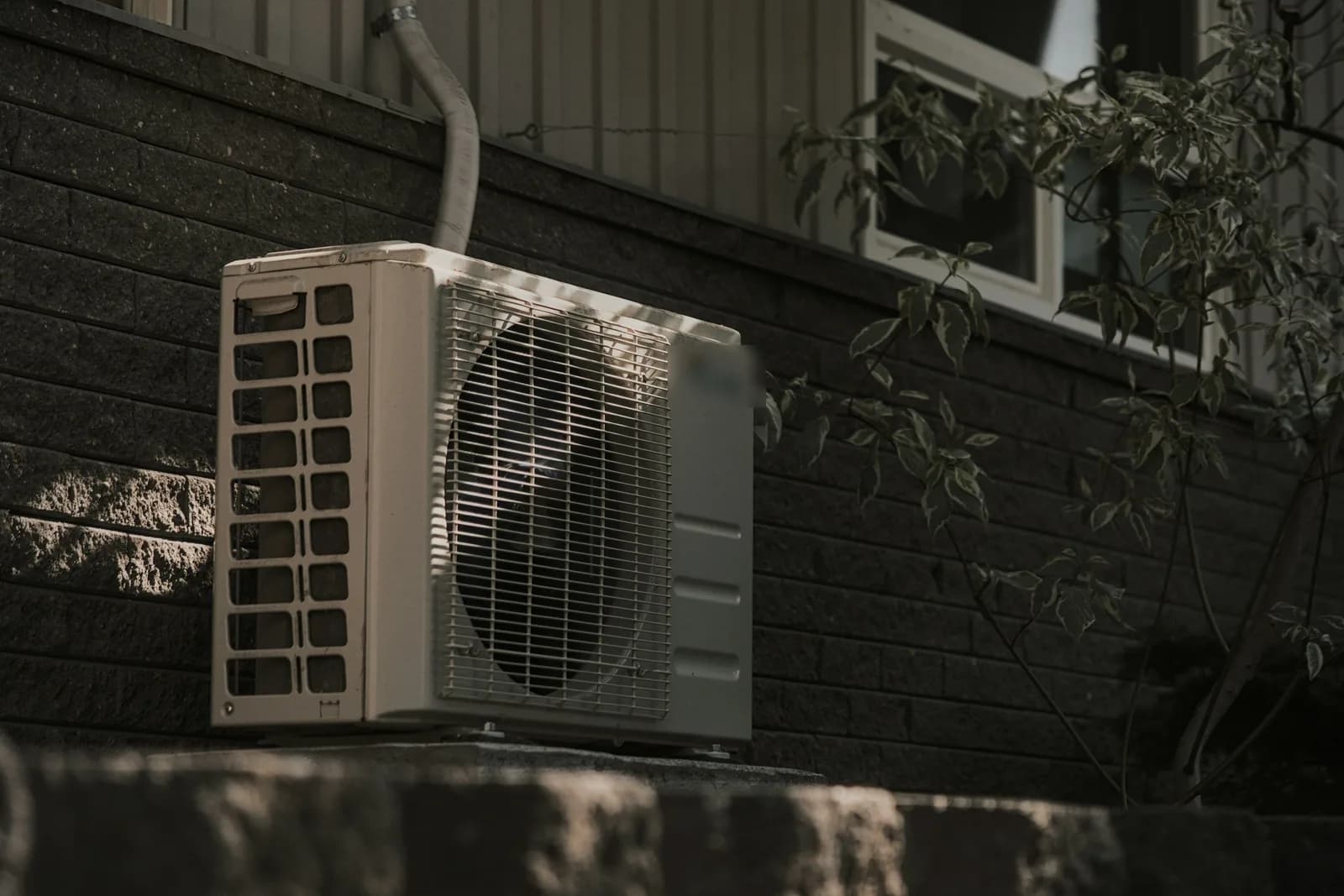 An outdoor central air conditioning condenser unit mounted on a stone foundation wall beneath a house window, with greenery alongside.