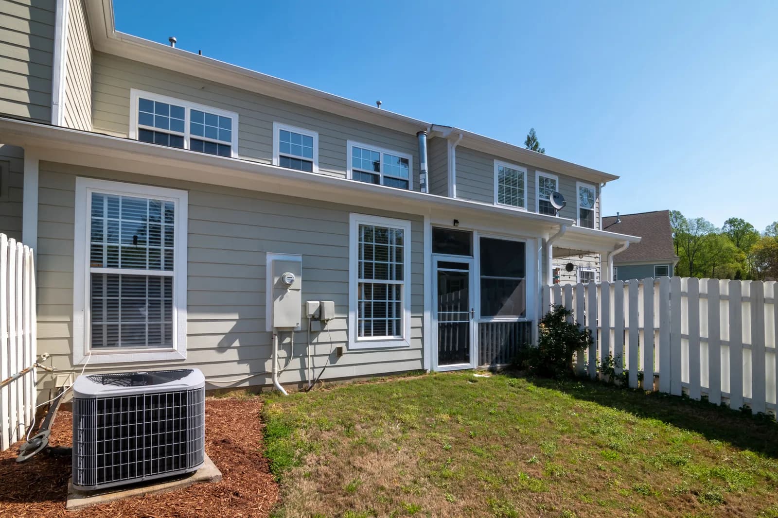 A round-top central air conditioning condenser unit in the mulched side yard of a vinyl-sided house with a white picket fence.