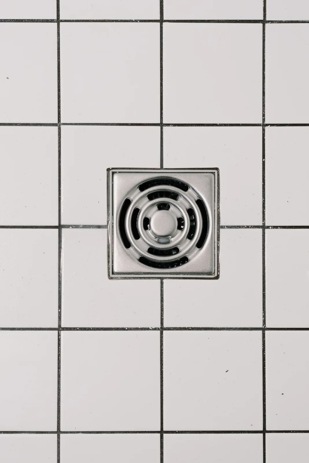 Overhead view of a square stainless steel shower floor drain cover set into white bathroom tile.