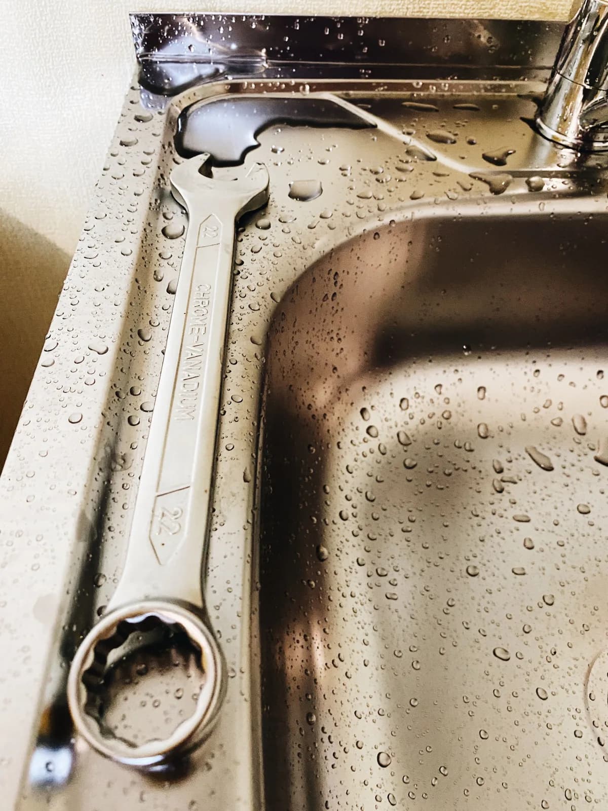 A combination wrench resting in a wet stainless steel kitchen sink basin next to the faucet, staged for a plumbing repair.