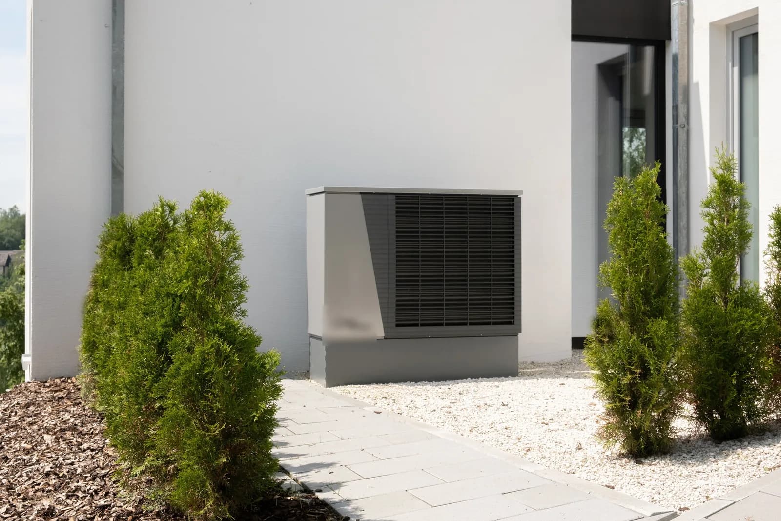 A heat pump condenser unit beside a modern white house entrance, flanked by trimmed evergreen shrubs and a gravel path.