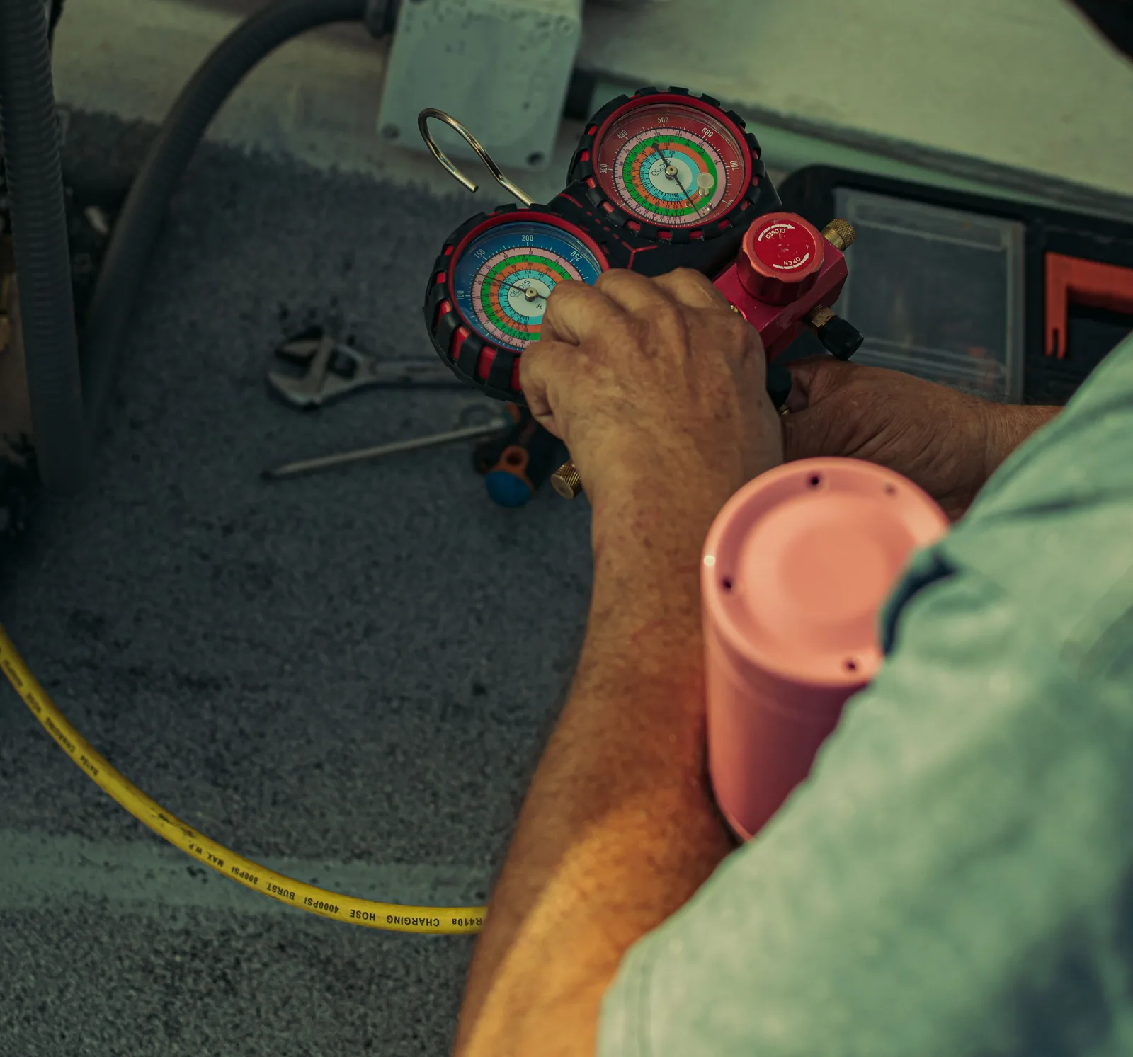 Close-up of a technician's hands operating a red-and-black refrigerant manifold gauge set connected to a yellow charging hose, with tools on the ground nearby.