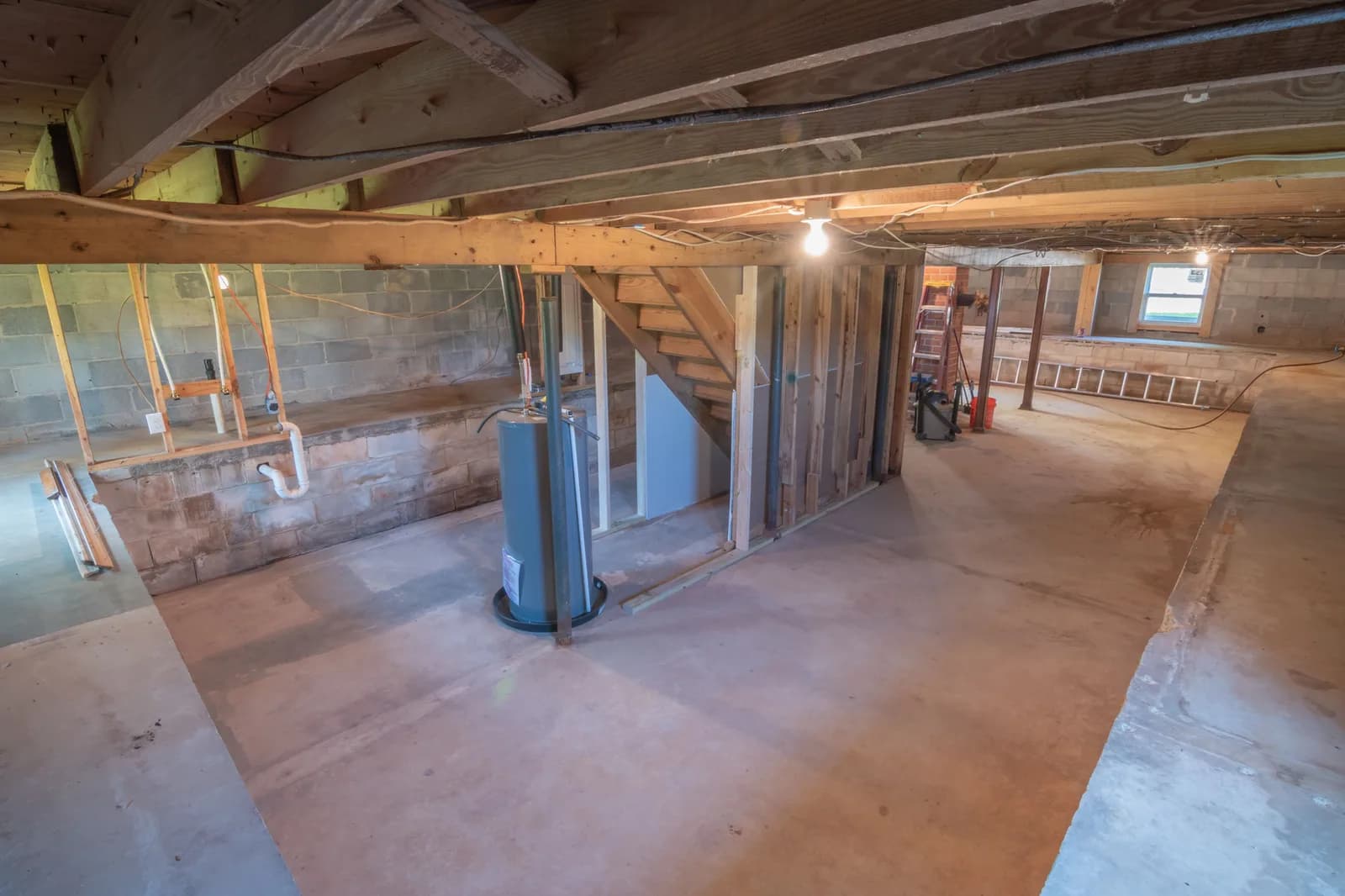 Unfinished basement of a house under construction, with exposed wood floor joists, cinderblock foundation walls, a new water heater, visible pipe fittings, and a framed stairway enclosure.