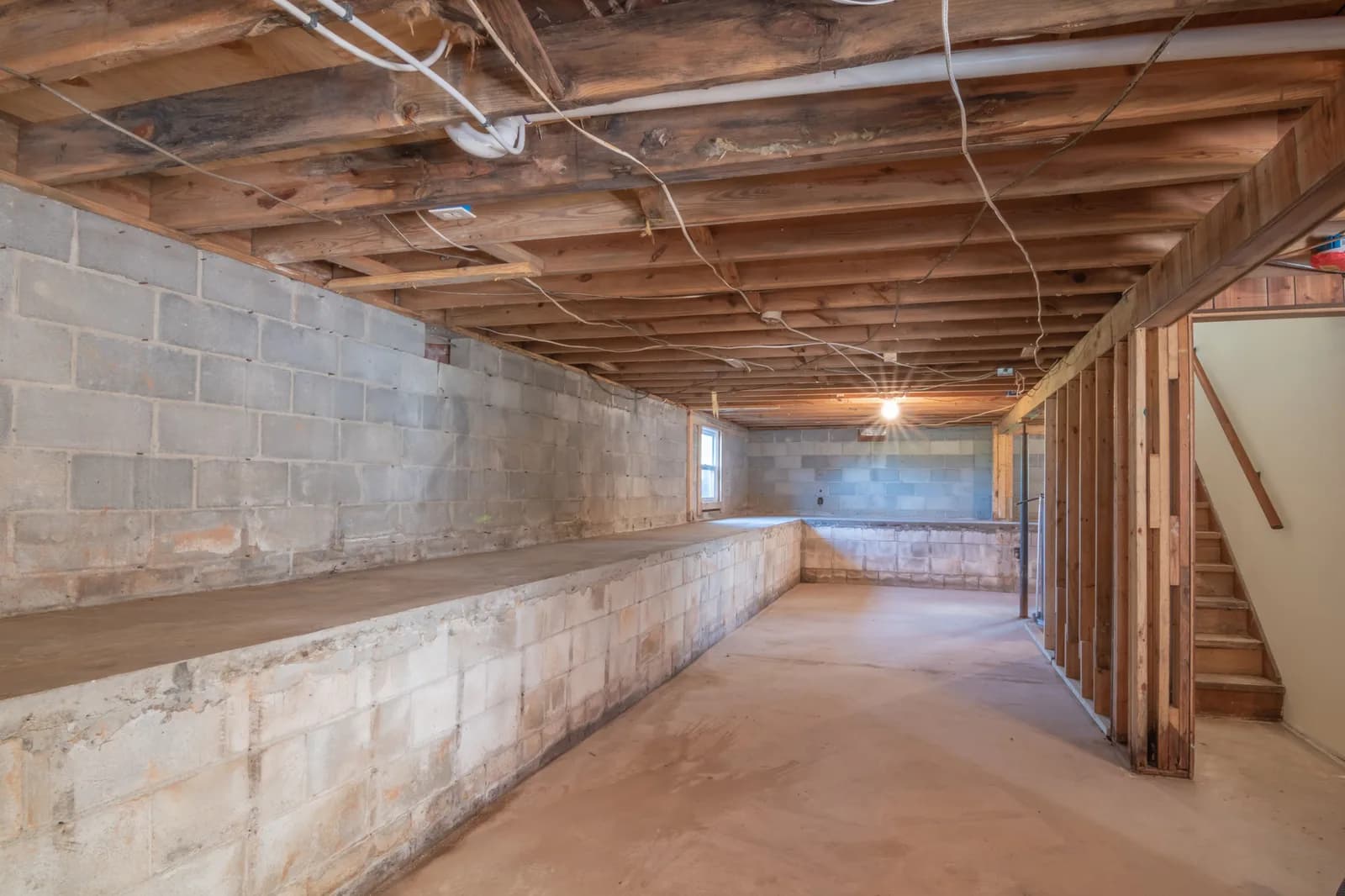 Unfinished house basement with cinderblock foundation walls and exposed wood ceiling joists, a white PVC pipe running along the joist bay, and a wood-stud partition wall framing a stairway before drywall.