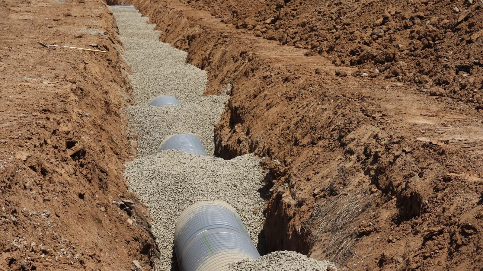 A close-up view down an open earthen trench showing a large corrugated pipe being bedded in gravel for underground pipe installation.