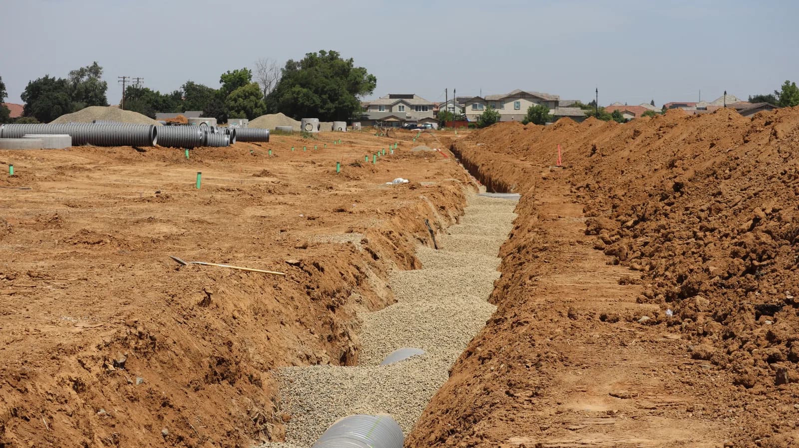 A wide view of an open excavation trench running through a residential subdivision, with stacked pipe sections and houses visible in the background.