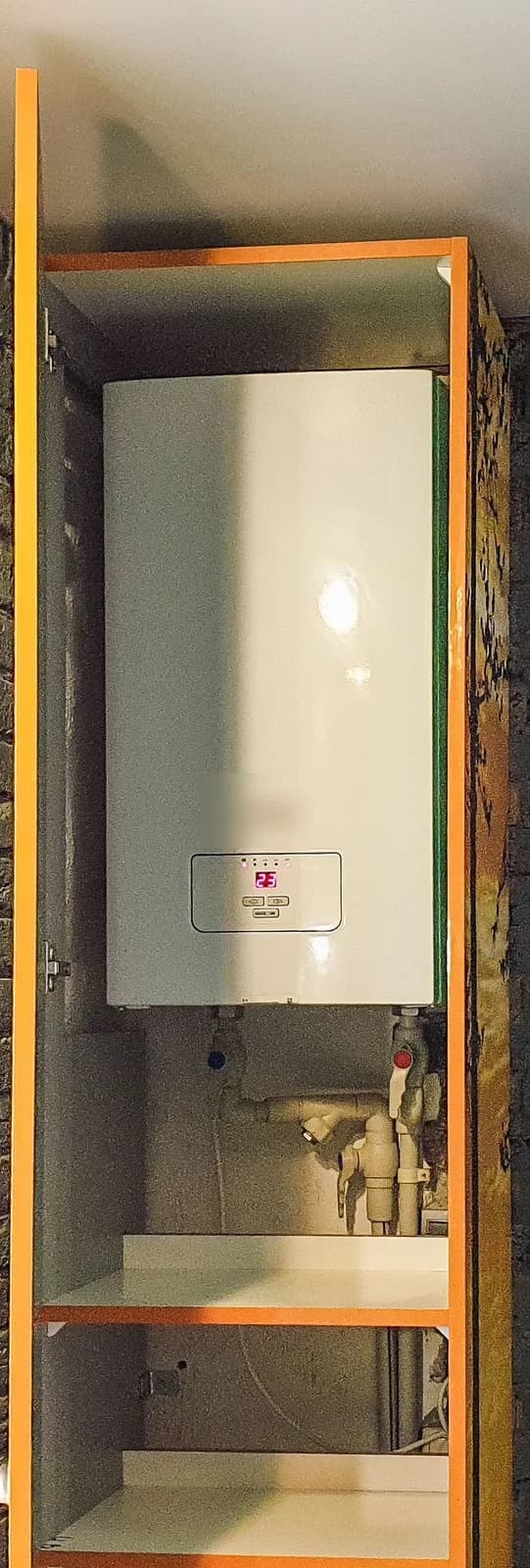 A white wall-mounted tankless combi boiler installed in an open utility closet, showing its digital temperature display and pipe connections below.