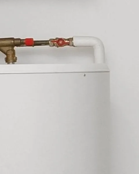 Close-up of a brass shutoff valve, tee fitting, and PVC pipe transition on top of a white residential water heater tank against a plain wall.