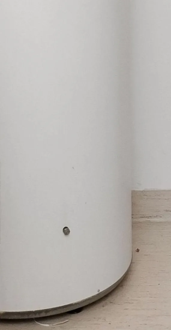 The lower curved body of a white residential water heater tank with an access panel, standing on a wood-look floor.