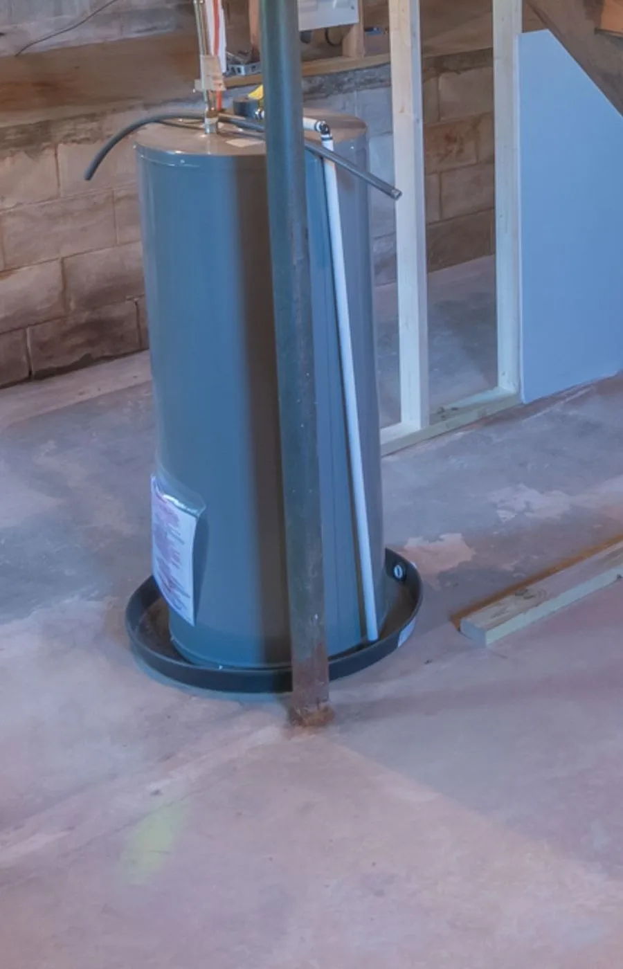 A gray cylindrical residential tank-style water heater with a drain pan, standing in an unfinished basement with a cinderblock wall.